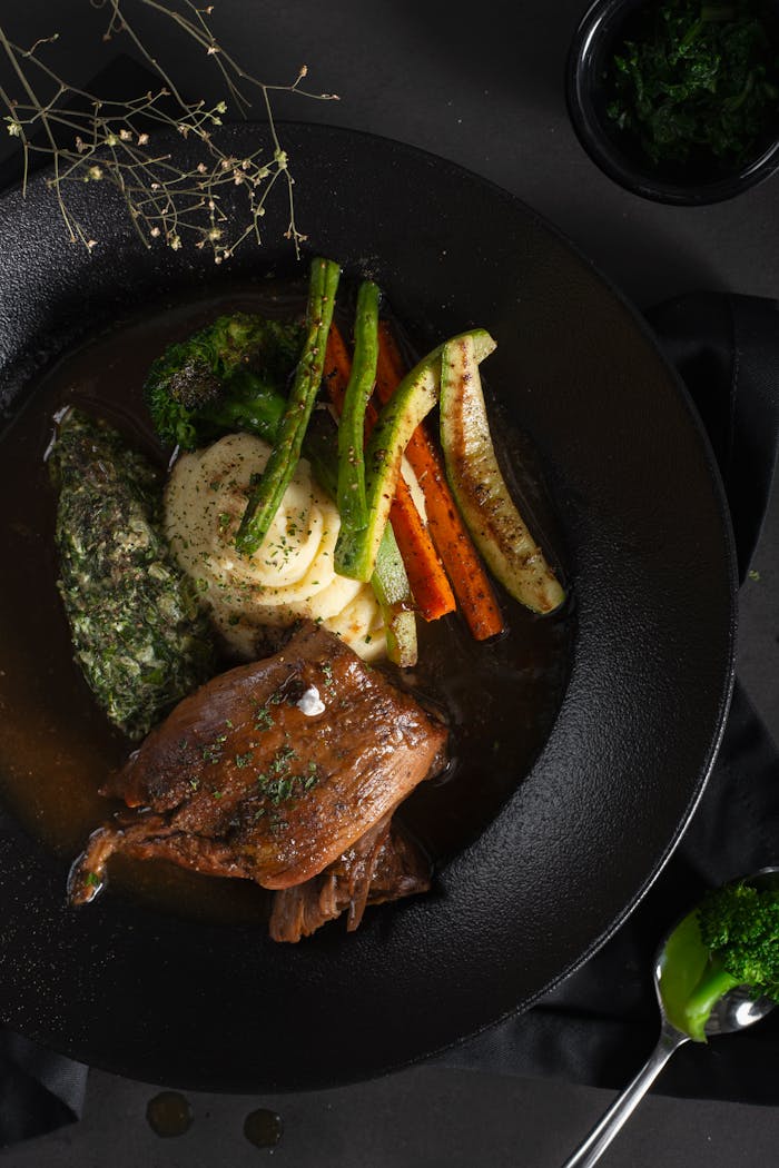 Elegant gourmet dish featuring braised meat with a side of vibrant vegetables and herbs served on a black plate.